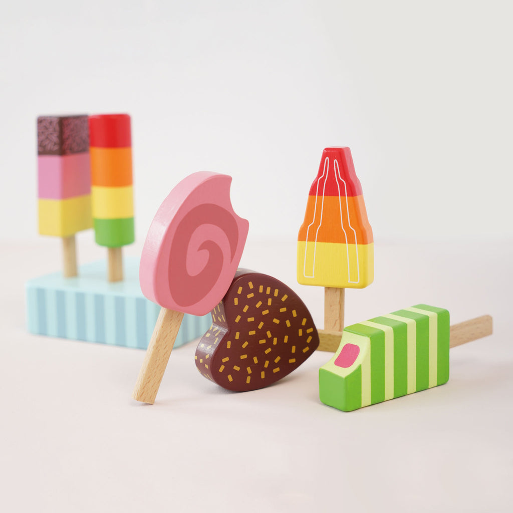 Six colorful wooden toy ice creams on sticks are arranged on a striped stand. They vary in shapes and vibrant designs, exuding a playful and whimsical tone.