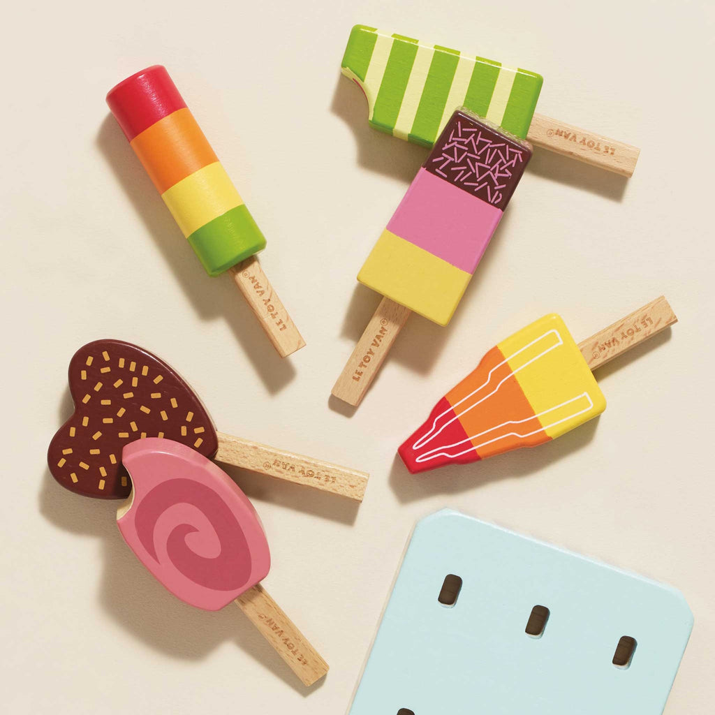 Six colorful wooden toy ice creams on sticks are arranged on a striped stand. They vary in shapes and vibrant designs, exuding a playful and whimsical tone.