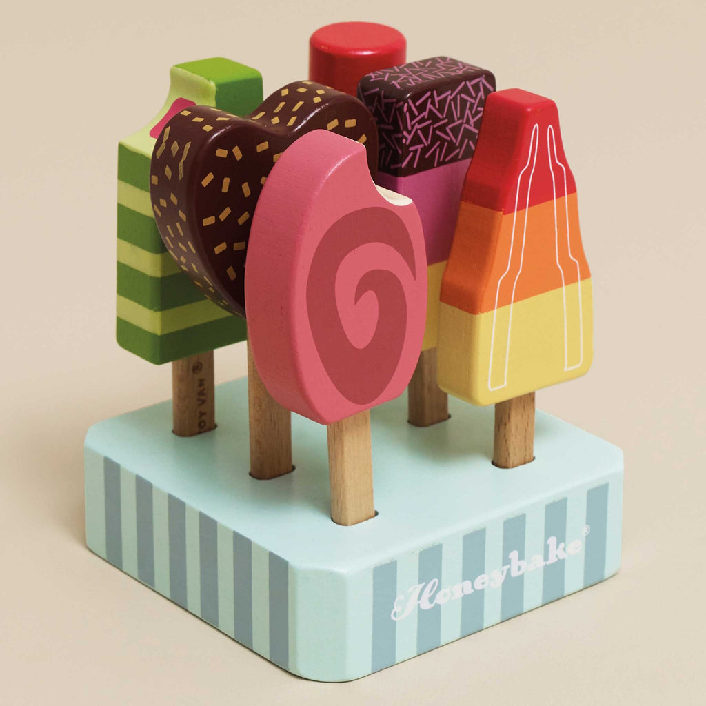 Six colorful wooden toy ice creams on sticks are arranged on a striped stand. They vary in shapes and vibrant designs, exuding a playful and whimsical tone.