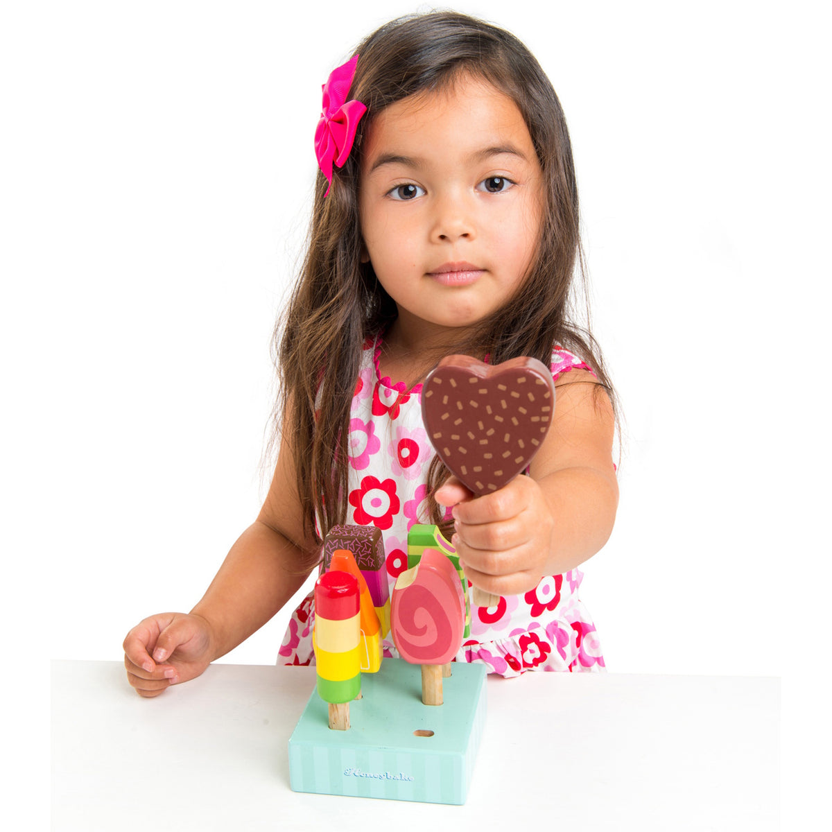A young girl wearing a floral dress holding up a chocolate heart-shaped wooden play popsicle, with the set's stand in front of her.