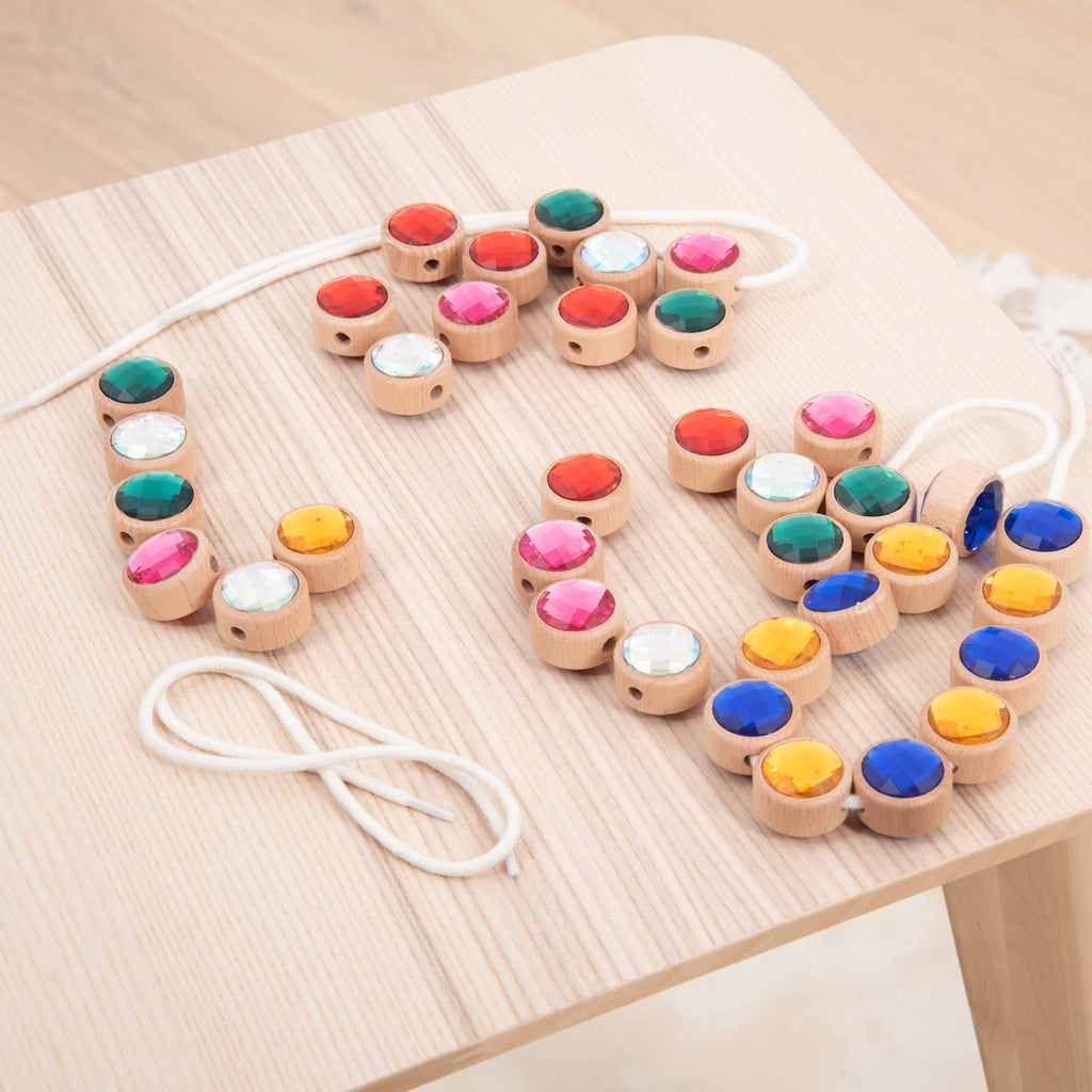 Wooden beads with colorful gems are arranged on a light wooden table. A white string with a needle is nearby, ready for threading.