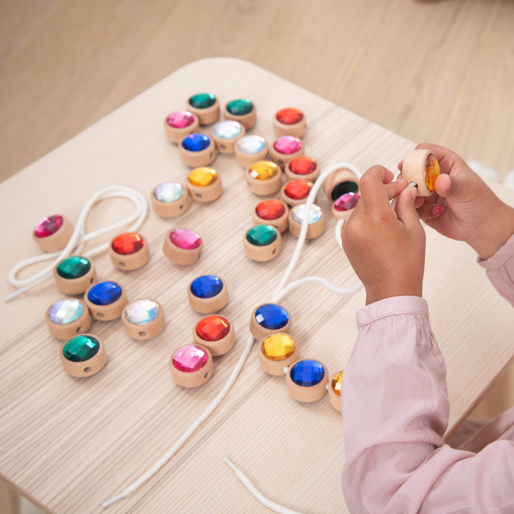 Wooden beads with colorful gems are arranged on a light wooden table. A white string with a needle is nearby, ready for threading.