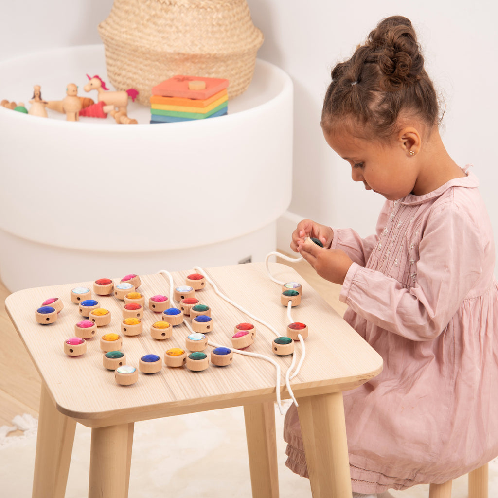 A young child in a pink dress focused on threading colorful wooden beads on strings at a small table. The scene conveys concentration and playfulness.