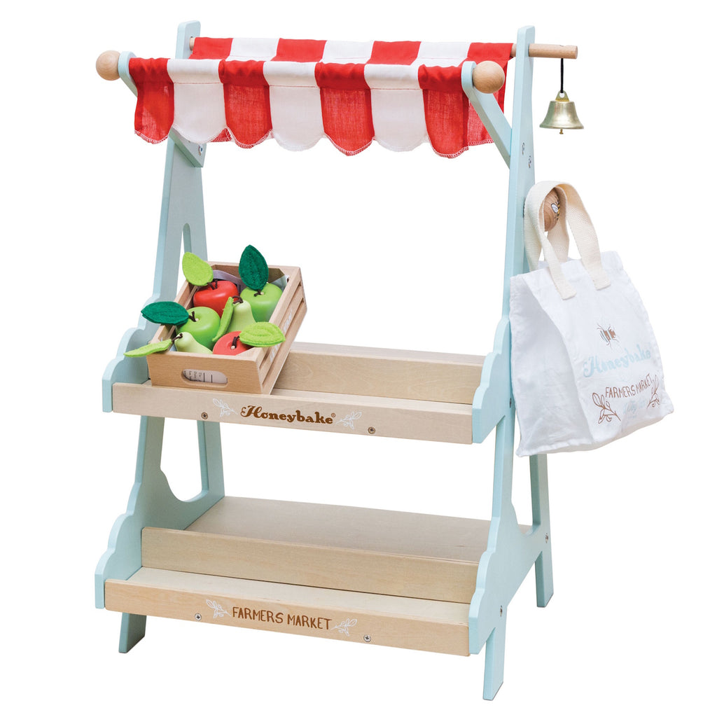 Toy market stall with red and white striped canopy and a bell. Features wooden tiers, a crate with toy fruits, and a canvas shopping bag labeled "Farmers Market."