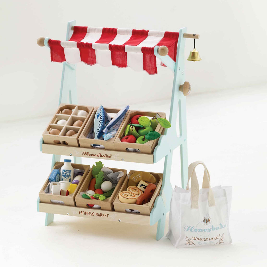 Toy market stall with red and white striped canopy, wooden trays of play groceries like eggs, fish, and vegetables, and a small white bag beside it.