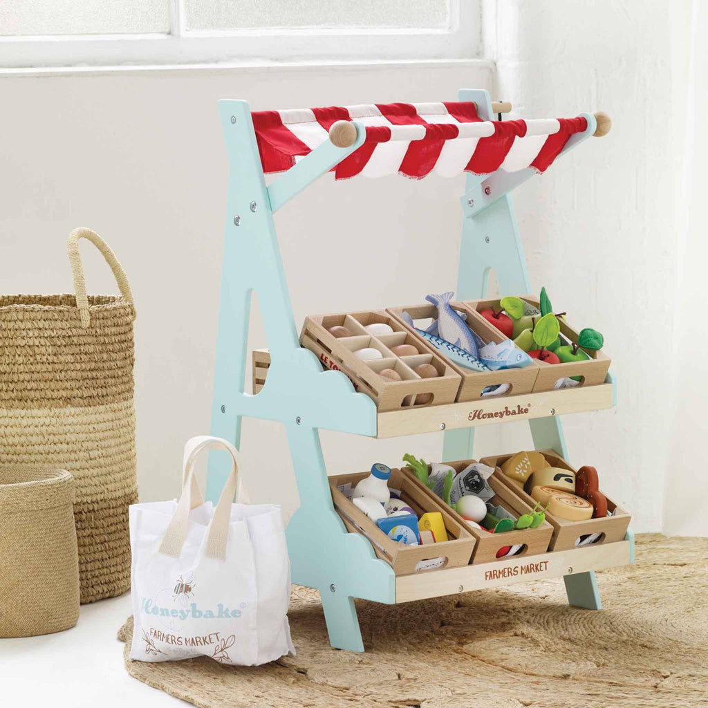 Toy market stall with red and white striped canopy, wooden trays of play groceries like eggs, fish, and vegetables, and a small white bag beside it.
