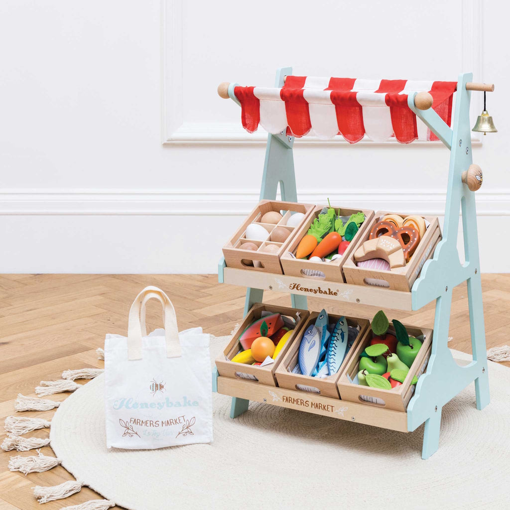 Toy market stall with red and white striped canopy, wooden trays of play groceries like eggs, fish, and vegetables, and a small white bag beside it.