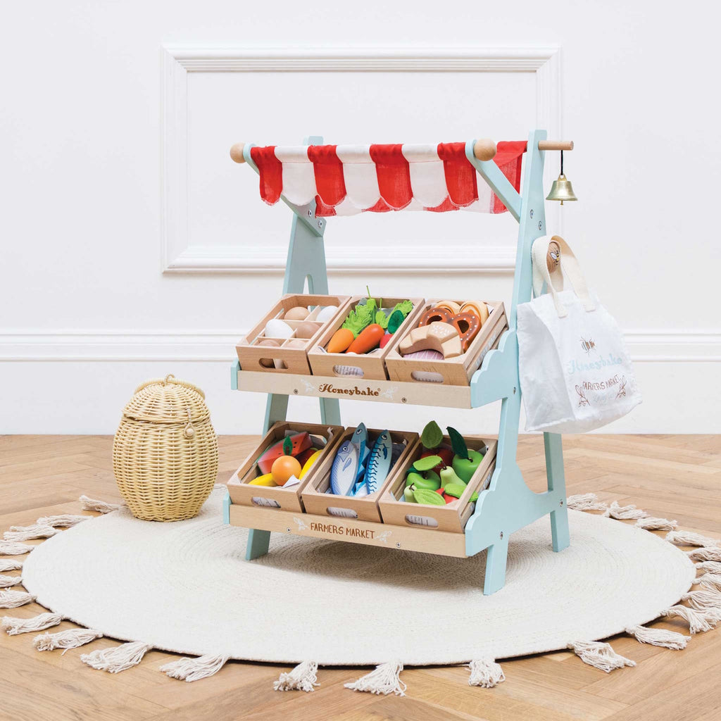 Toy market stall with red and white striped canopy, wooden trays of play groceries like eggs, fish, and vegetables, and a small white bag beside it.