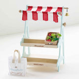 Wooden toy market stall with red striped awning, crate of play fruit, bell, and shopping tote bag.
