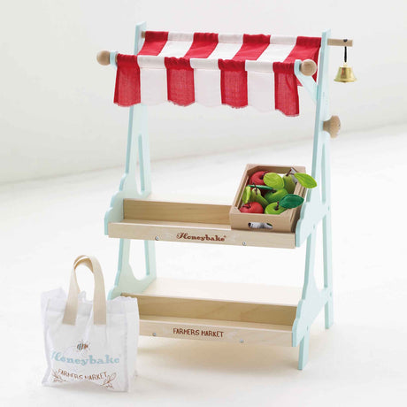 Wooden toy market stall with red striped awning, crate of play fruit, bell, and shopping tote bag.