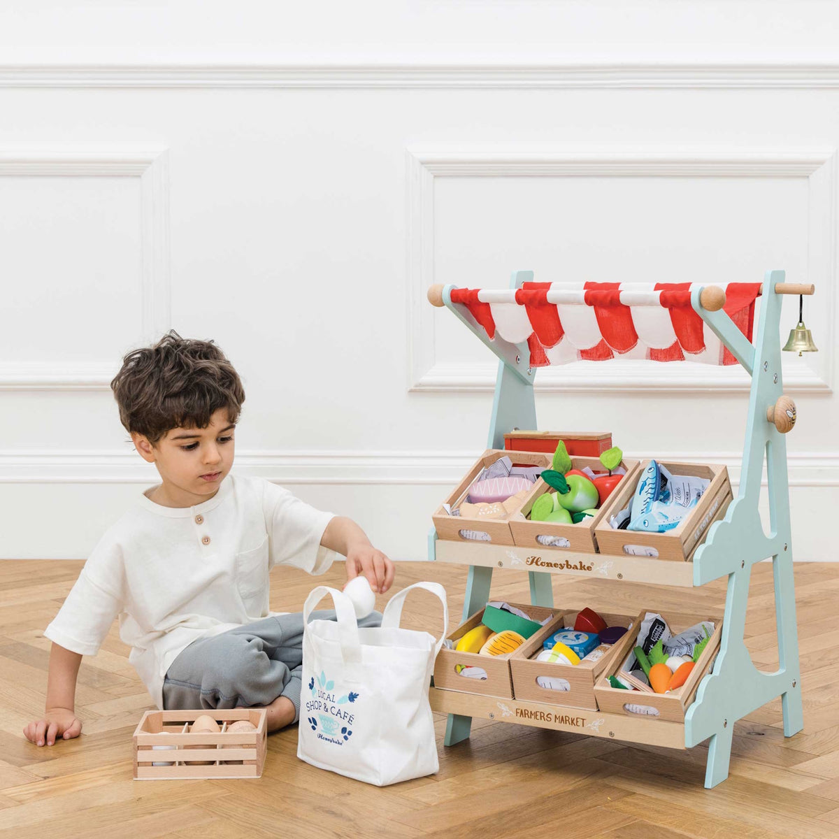 Child sitting next to a wooden market stall play set filled with toy food crates and tote bag.