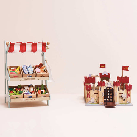 Wooden toy farmers market stall filled with play food crates next to a wooden toy castle.