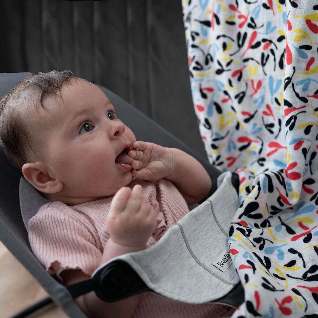 A baby wearing a pink outfit lies in a gray bouncer, gazing intently at a colorful patterned blanket. The scene conveys curiosity and wonder.