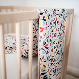 White sensory muslin cloth with a colorful leaf pattern draped over the railing of a wooden baby crib.