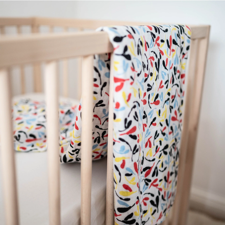 White sensory muslin cloth with a colorful leaf pattern draped over the railing of a wooden baby crib.
