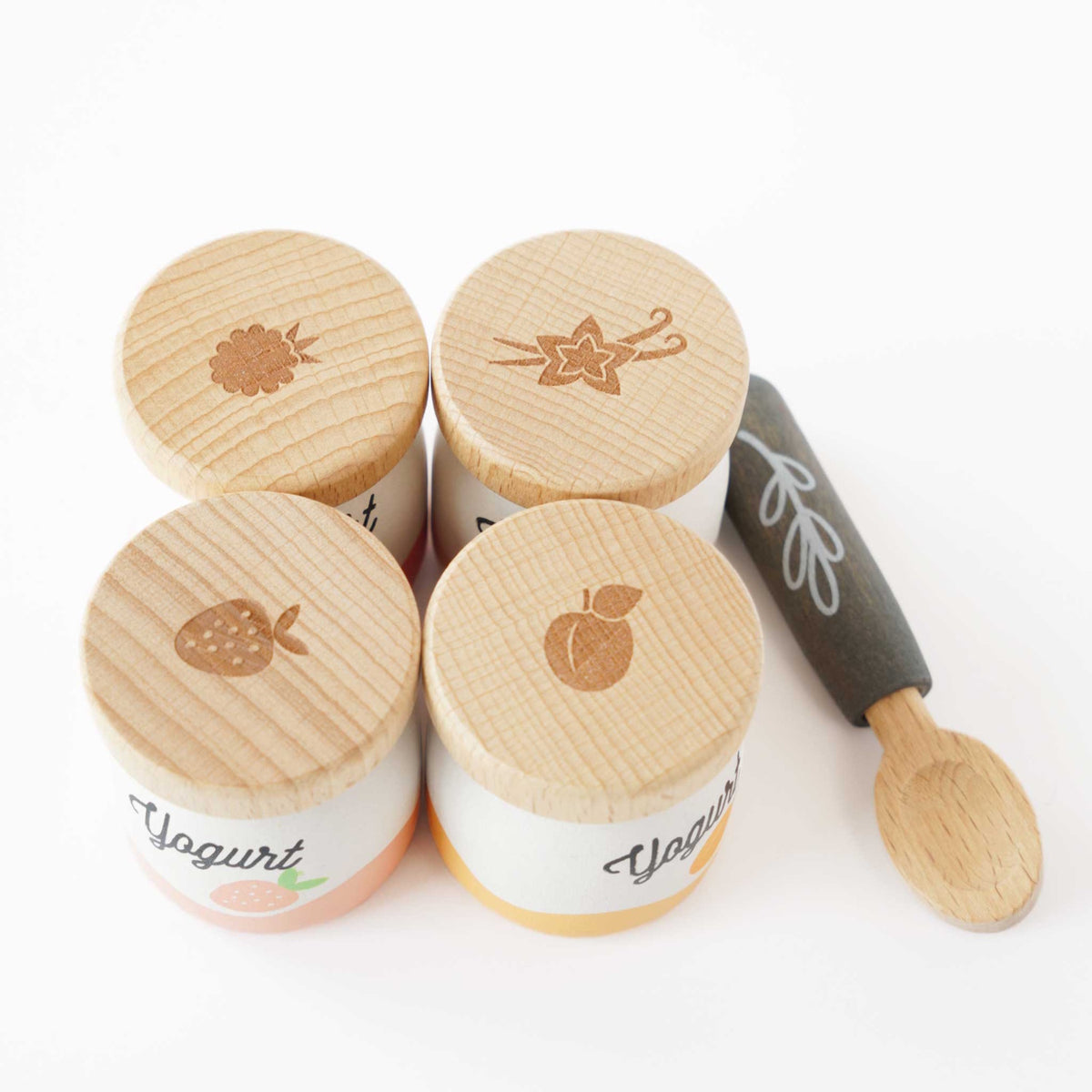 Top view of four wooden play yogurt pots with fruit-embossed lids next to a toy spoon.