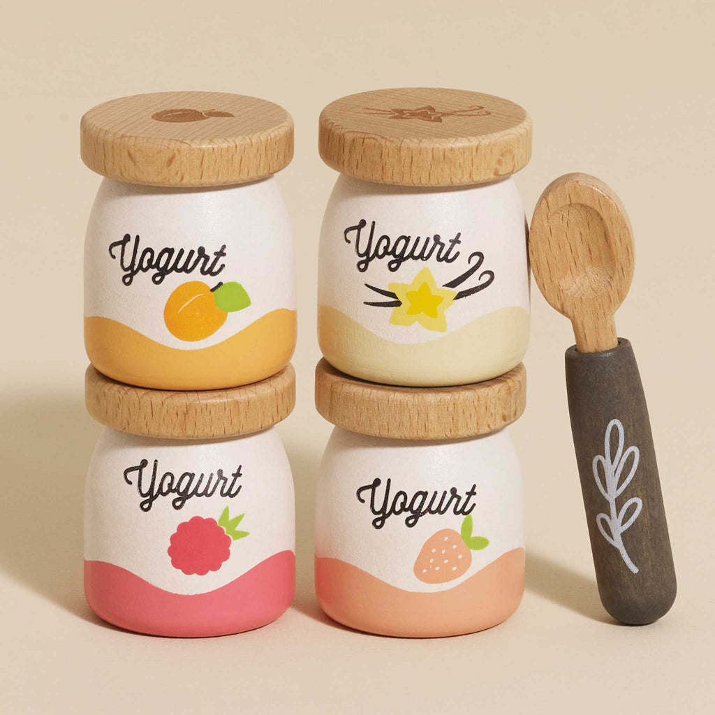 Four wooden yogurt jars with fruit illustrations—apricot, vanilla, raspberry, and strawberry—stacked in two columns, next to a wooden spoon.