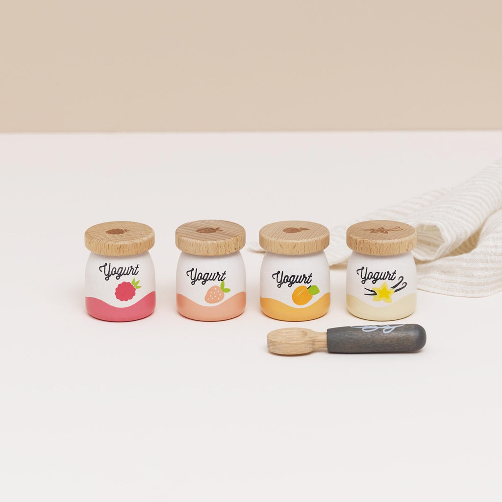 Four small wooden yogurt jars with fruit designs—raspberry, strawberry, peach, vanilla—each with a wooden lid. A wooden spoon lies nearby.