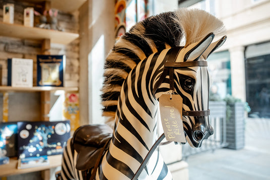 A zebra-themed rocking horse with bold black-and-cream stripes and a hanging tag that reads 'Please ride me,' photographed at My Small World Toy Store.