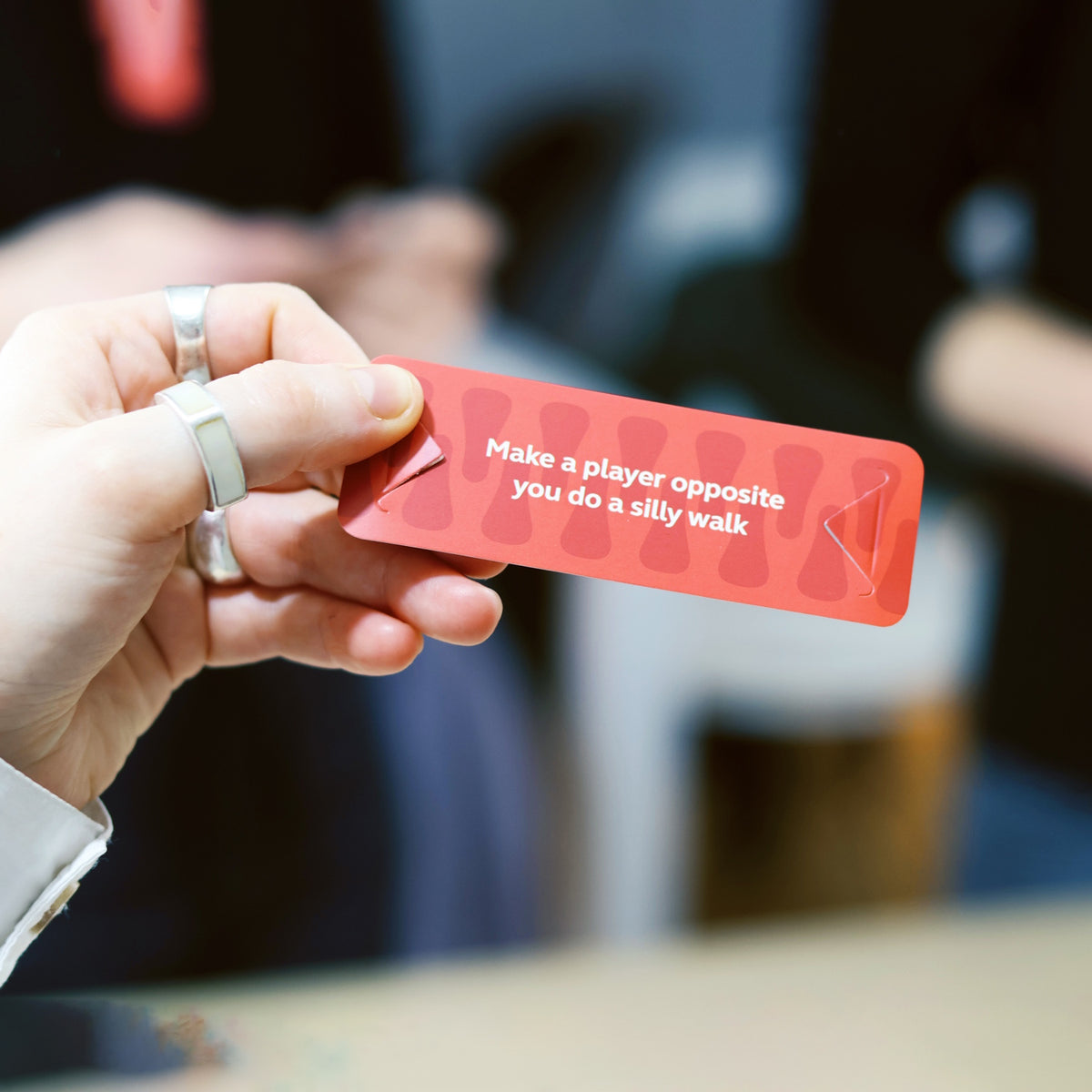 A hand holding a red game card that reads "Make a player opposite you do a silly walk".