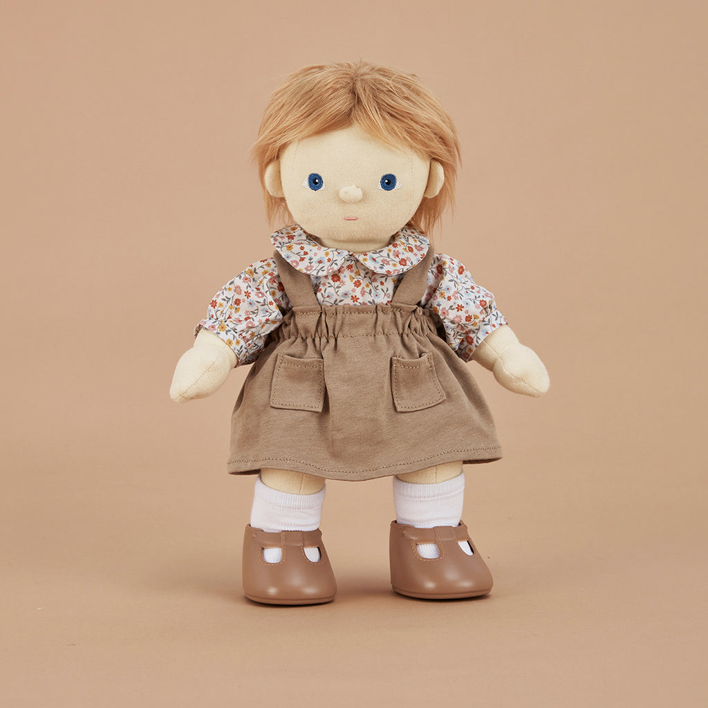 A picture showing dinkum doll poppet wearing the dinkum doll clothing set prairie dress outfit - a floral blouse, brown pinafore dress white socks and brown shoes in front of a beige background