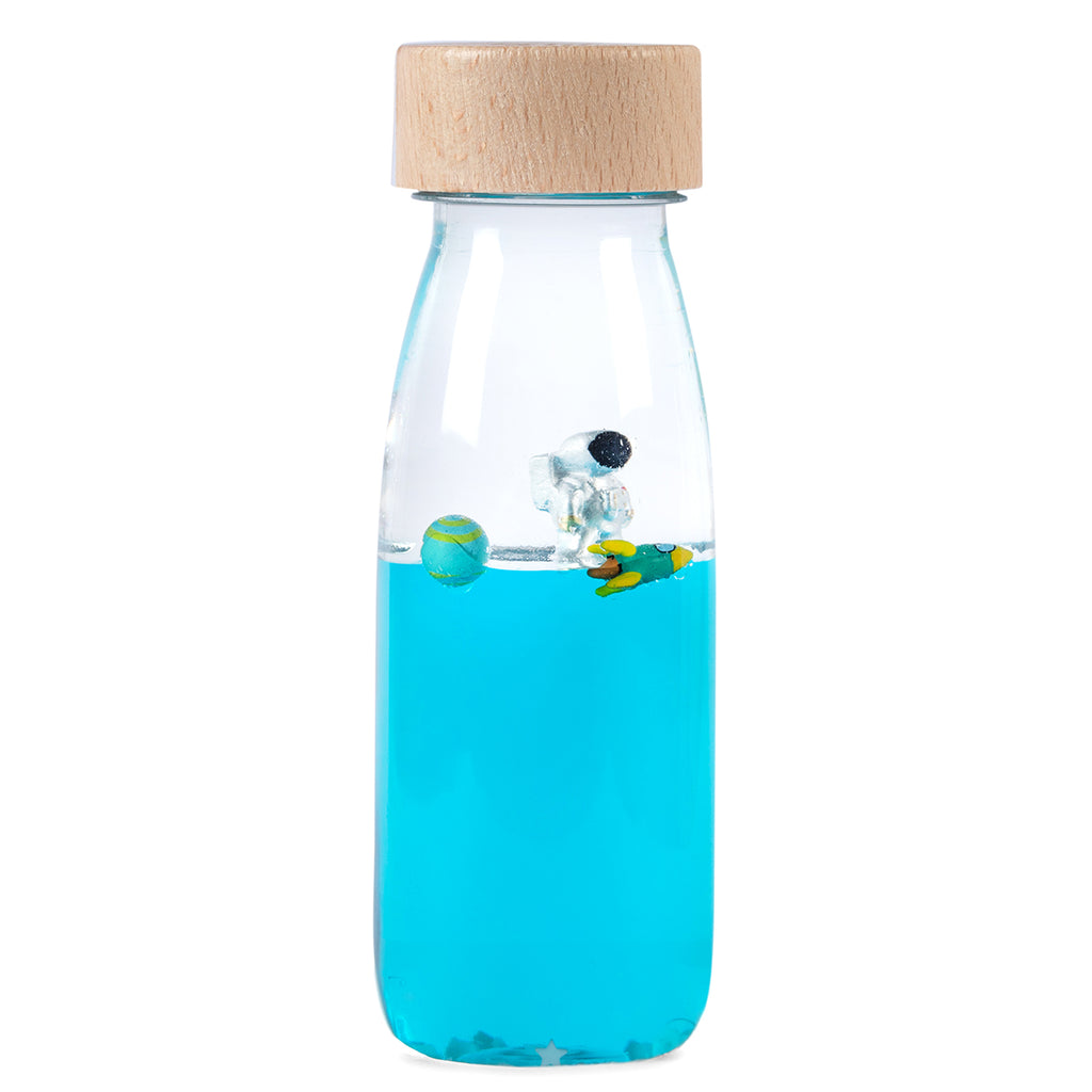 A clear sensory bottle with a wooden cap, filled with blue liquid, a floating astronaut figurine, and silver star-shaped confetti.